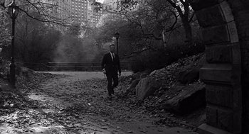 Movie still from “Mirage” (1965), directed by Edward Dmytryk – A black and white photo of a man in a suit walking in a park; Wide shot, Over the shoulder angle