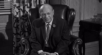 Movie still from “Mirage” (1965), directed by Edward Dmytryk – An older man sitting in a chair wearing a suit and tie; Medium shot, High angle