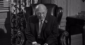 Movie still from “Mirage” (1965), directed by Edward Dmytryk – An older man sitting in a chair wearing a suit and tie; Medium shot, High angle