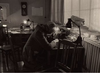 Movie still from “Mirror” (1975), directed by Andrei Tarkovsky – An older woman sitting at a desk taking a picture of papers; Medium shot, Over the shoulder angle
