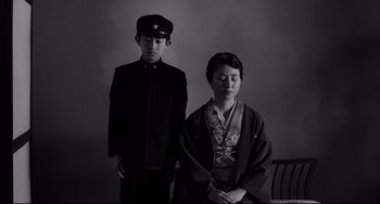 Movie still from “Mishima: A Life in Four Chapters” (1985), directed by Paul Schrader – A man and a woman in traditional japanese garb stand side by side in front of a wall; Medium shot, Low angle