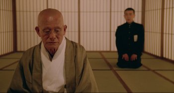 Movie still from “Mishima: A Life in Four Chapters” (1985), directed by Paul Schrader – A man sitting on the ground in front of another man; Medium shot, Low angle