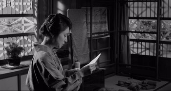 Movie still from “Mishima: A Life in Four Chapters” (1985), directed by Paul Schrader – A woman sitting in a chair reading a paper; Medium shot, Over the shoulder angle