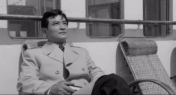 Movie still from “Mishima: A Life in Four Chapters” (1985), directed by Paul Schrader – A black and white photo of a man sitting on a bench; Medium shot, Low angle