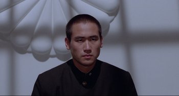 Movie still from “Mishima: A Life in Four Chapters” (1985), directed by Paul Schrader – A man in a black shirt is in front of a white wall; Close Up shot, Low angle