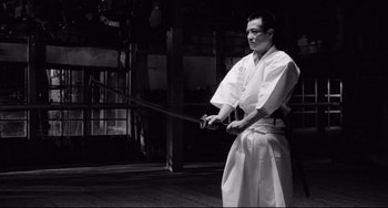 Movie still from “Mishima: A Life in Four Chapters” (1985), directed by Paul Schrader – A man holding a sword while standing on a wooden floor; Medium shot, Low angle
