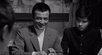 Movie still from “Mishima: A Life in Four Chapters” (1985), directed by Paul Schrader – A man wearing a suit and tie smiling for the camera; Close Up shot, Over the shoulder angle