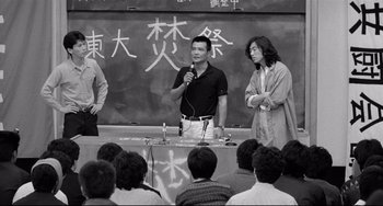 Movie still from “Mishima: A Life in Four Chapters” (1985), directed by Paul Schrader – A man standing in front of a group of people in front of a chalkboard; Medium shot, Over the shoulder angle