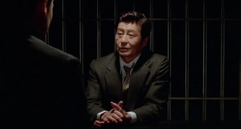 Movie still from “Mishima: A Life in Four Chapters” (1985), directed by Paul Schrader – A man wearing a suit and tie sitting in front of a wall; Close Up shot, Over the shoulder angle