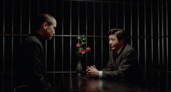 Movie still from “Mishima: A Life in Four Chapters” (1985), directed by Paul Schrader – Two men sitting at a table in front of a vase of flowers; Medium shot, High angle