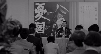 Movie still from “Mishima: A Life in Four Chapters” (1985), directed by Paul Schrader – A man is speaking at an event in front of a poster of an officer; Medium shot, Low angle