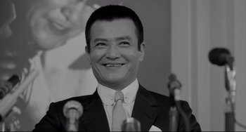 Movie still from “Mishima: A Life in Four Chapters” (1985), directed by Paul Schrader – A black and white photo of a man in a business suit; Close Up shot, Low angle