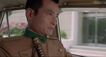 Movie still from “Mishima: A Life in Four Chapters” (1985), directed by Paul Schrader – A man in a tan jacket and a green neck tie; Close Up shot, Low angle
