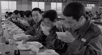 Movie still from “Mishima: A Life in Four Chapters” (1985), directed by Paul Schrader – A black and white photo of a group of people eating; Medium shot, Low angle