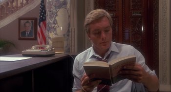 Movie still from “Missing” (1982), directed by Costa-Gavras – A man sitting at a desk reading a book in front of an american flag; Medium shot, Low angle