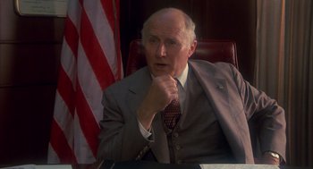 Movie still from “Missing” (1982), directed by Costa-Gavras – An older man sitting in a chair in front of an american flag; Close Up shot, High angle