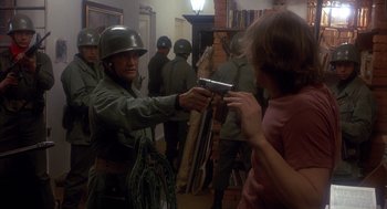 Movie still from “Missing” (1982), directed by Costa-Gavras – A group of people in a room with a man holding a gun; Medium shot, Over the shoulder angle