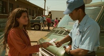 Movie still from “Mississippi Masala” (1991), directed by Mira Nair – A man in a blue hat is handing a card to a woman in an orange jacket; Medium shot, Over the shoulder angle