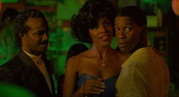 Movie still from “Mississippi Masala” (1991), directed by Mira Nair – Two men and a woman posing for a picture in front of a green wall; Close Up shot, Over the shoulder angle