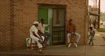 Movie still from “Mississippi Masala” (1991), directed by Mira Nair – A group of men sitting on stools in front of a building; Wide shot, Low angle