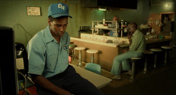 Movie still from “Mississippi Masala” (1991), directed by Mira Nair – A man sitting at a table in front of a counter; Medium shot, Over the shoulder angle