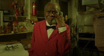 Movie still from “Mississippi Masala” (1991), directed by Mira Nair – An older man wearing a red jacket holding a phone to his ear; Medium shot, High angle