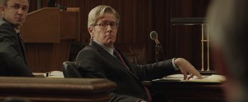 Movie still from “Molly's Game” (2017), directed by Aaron Sorkin – A man sitting in a chair in front of a microphone in a courtroom; Medium shot, Over the shoulder angle