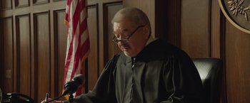Movie still from “Molly's Game” (2017), directed by Aaron Sorkin – An older man in a robe and glasses is speaking into a microphone in front of the american flag; Close Up shot, Low angle