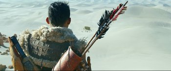 Movie still from “Monster Hunter” (2020), directed by Paul W.S. Anderson – A young boy holding a bow and arrow in the sand; Close Up shot, Over the shoulder angle