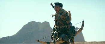Movie still from “Monster Hunter” (2020), directed by Paul W.S. Anderson – A man in native american garb holding a bow and arrow; Medium shot, Over the shoulder angle