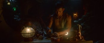 Movie still from “Monster Hunter” (2020), directed by Paul W.S. Anderson – A man is cutting food on a table in the dark; Medium shot, Low angle