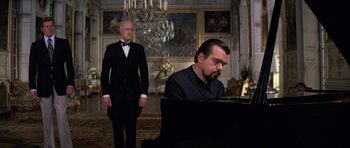 Movie still from “Moonraker” (1979), directed by Lewis Gilbert – A man in a suit is playing piano in front of another man in a suit; Medium shot, Low angle