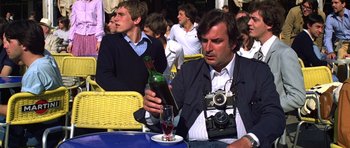 Movie still from “Moonraker” (1979), directed by Lewis Gilbert – A man sitting at an outdoor table with a wine bottle and a camera; Close Up shot, Low angle