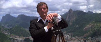 Movie still from “Moonraker” (1979), directed by Lewis Gilbert – A man in a black suit is holding a camera on a tripod; Medium shot, Low angle