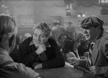 Movie still from “Moontide” (1942), directed by Fritz Lang – A man sitting at a table with a woman in the background; Medium shot, Over the shoulder angle