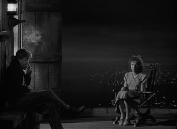 Movie still from “Moontide” (1942), directed by Fritz Lang – A man and a woman sitting on a chair in front of a building; Wide shot, High angle