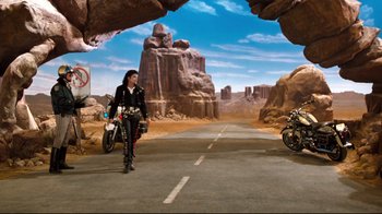 Movie still from “Moonwalker” (1988), directed by Jerry Kramer – A woman walking down a road with a motorcycle; Wide shot, High angle