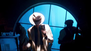 Movie still from “Moonwalker” (1988), directed by Jerry Kramer – A man wearing a white hat holding a microphone in front of a window; Medium shot, High angle
