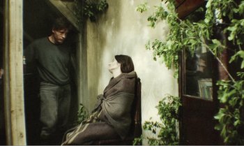 Movie still from “Mother and Son” (1997), directed by Aleksandr Sokurov – A man standing next to a woman sitting on a chair; Wide shot, High angle