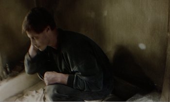 Movie still from “Mother and Son” (1997), directed by Aleksandr Sokurov – A man sitting on the ground with a cigarette in his mouth; Medium shot, High angle