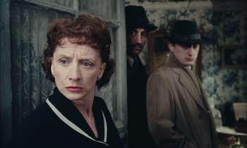 Movie still from “Mr. Klein” (1976), directed by Joseph Losey – A woman standing next to two men wearing suits and hats; Close Up shot, Over the shoulder angle