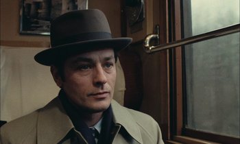 Movie still from “Mr. Klein” (1976), directed by Joseph Losey – A man wearing a hat and a trench coat standing in front of a window; Close Up shot, High angle