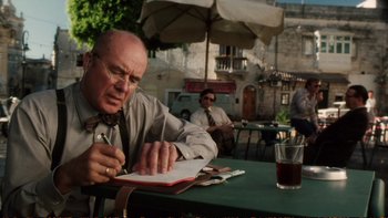 Movie still from “Munich” (2005), directed by Steven Spielberg – An older man sitting at an outdoor table writing in a notebook; Medium shot, Over the shoulder angle
