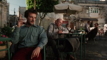 Movie still from “Munich” (2005), directed by Steven Spielberg – A man sitting at a table with another man sitting at a table with an umbrella; Medium shot, Low angle
