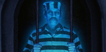 Movie still from “Muppets Haunted Mansion” (2021), directed by Kirk R. Thatcher – An old man wearing a striped shirt and a hat is standing behind bars; Medium shot, High angle