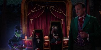 Movie still from “Muppets Haunted Mansion” (2021), directed by Kirk R. Thatcher – A couple of puppets sitting on top of coffins in front of a stage; Medium shot, Over the shoulder angle