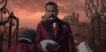 Movie still from “Muppets Haunted Mansion” (2021), directed by Kirk R. Thatcher – A man in a red suit holding a fan in front of a pink sky; Medium shot, Over the shoulder angle