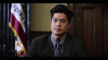 Movie still from “13 Reasons Why” (2017), created by Brian Yorkey – A man in a suit and tie is sitting at a podium; Close Up shot, Over the shoulder angle