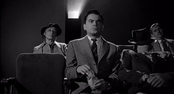 Movie still from “Murder by Contract” (1958), directed by Irving Lerner – A man in a suit and tie sitting next to an older woman; Medium shot, Low angle
