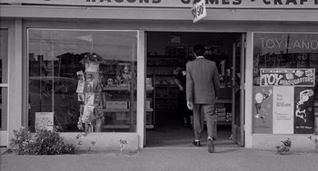 Movie still from “Murder by Contract” (1958), directed by Irving Lerner – A black and white photo of a man walking in front of a store; Wide shot, Over the shoulder angle